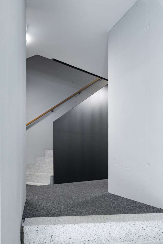 Podest im Treppenhaus: Sichtbeton, schwarze Stahlbrüstung und Teppichbelag in einer reduzierten Palette.