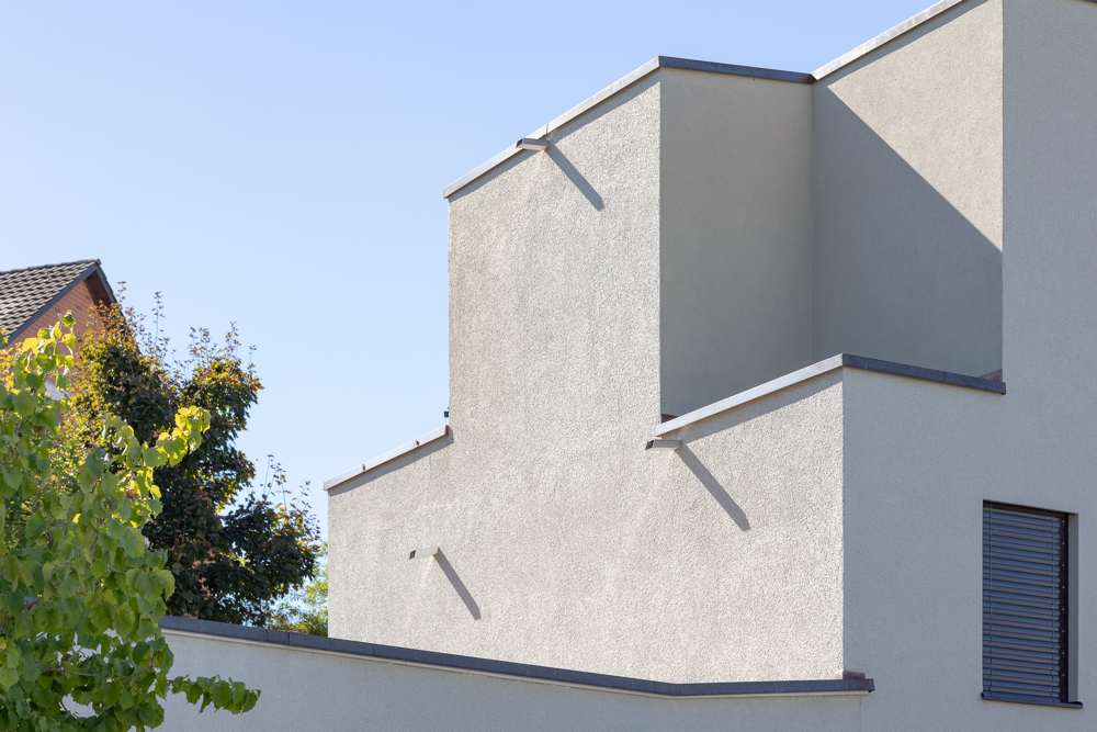 Gestaffelte Kuben der Attikageschosse im Gegenlicht: eingefärbter Abrieb auf Wärmedämmverbundsystem, Dachrandabschlüsse und Speier in Metall, Rafflamellenstoren vor den Öffnungen.