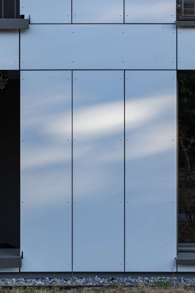 Plattenfassade im Streiflicht: grossformatige Faserzementplatten, sichtbar verschraubt, Fenster bündig in der Ebene.