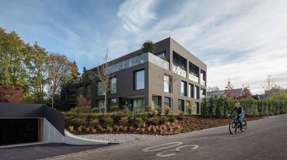 Ansicht von Nordwesten. Die hinterlüftete Fassade aus horizontalen, eloxierten Aluminiumprofilen läuft ohne Dachrandabschluss über Dachkante, Brüstung und Fläche hinweg; links die Einfahrt zur Tiefgarage.