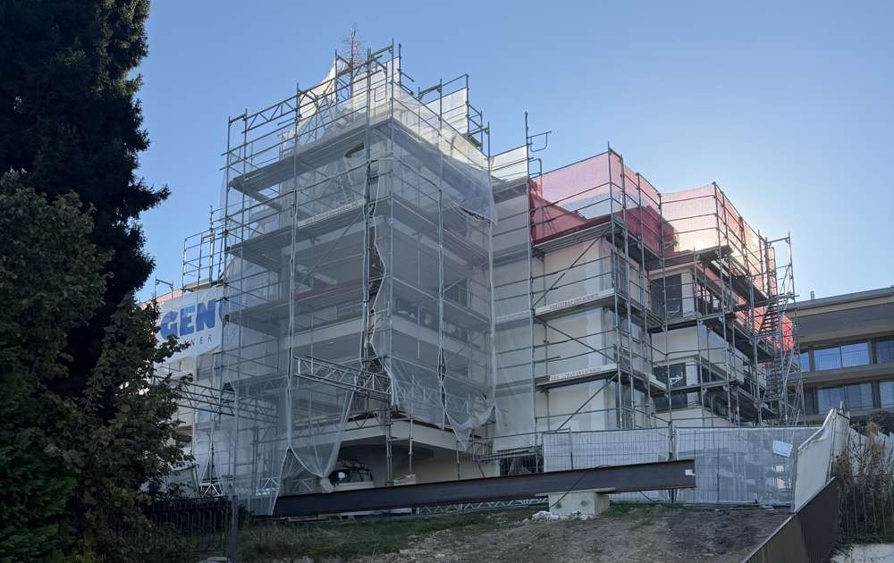 Baustelle im Oktober 2026: der Rohbau am Hang eingerüstet, das Steildach im Aufbau; davor die Böschung zur Baumschutzzone am Rand der Parzelle.