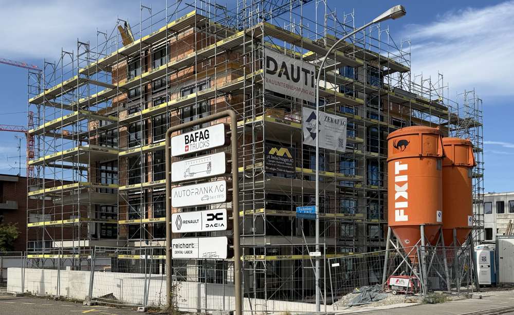 Baustelle an der Trockenloostrasse, September 2026: der Rohbau in Backstein und Stahlbeton im Gerüst, die Fensteröffnungen gesetzt; davor die Kranbahn und die Silos des Baumeisters.