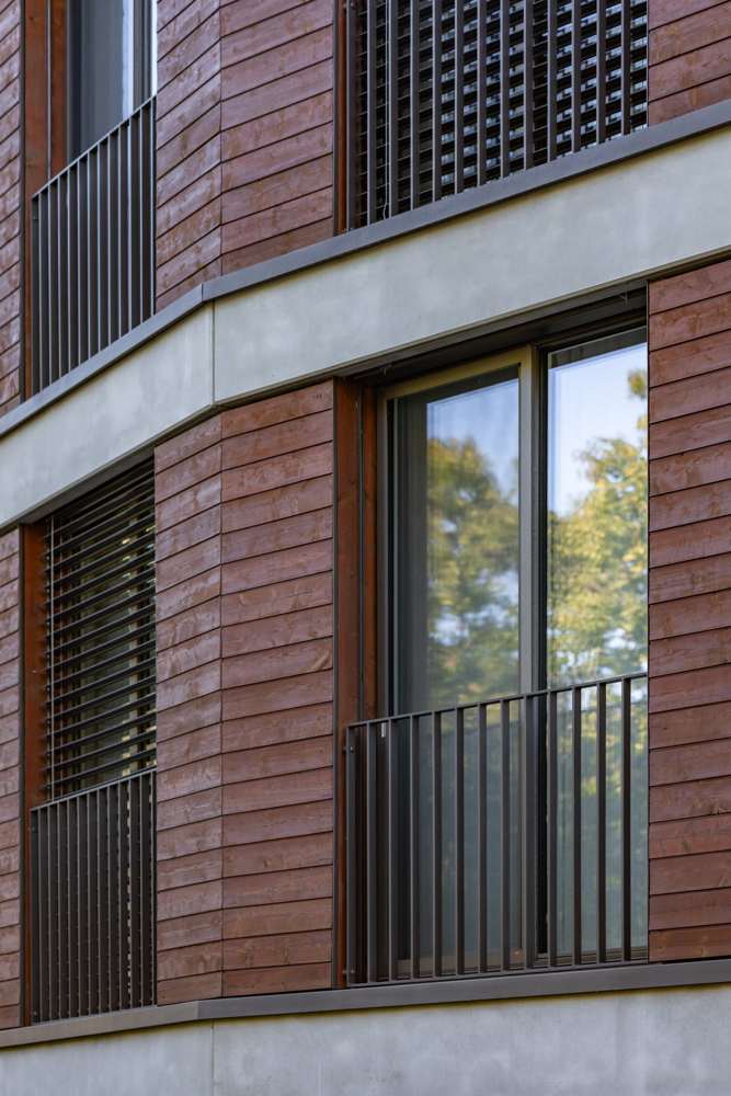 Fassadendetail: Betonbrüstungsband, Holzschalung, Holz-Metall-Fenster und Stabgeländer an der Gebäudekante.