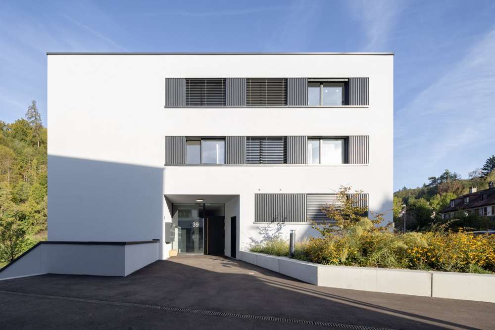 Haus 39 von der Zufahrt: Der Hauseingang liegt als Einschnitt im Baukörper, darüber die horizontalen Fensterbänder mit vertikal profilierten Metallpaneelen und Lamellenstoren.