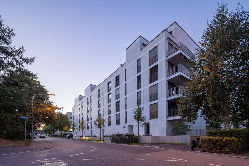 Abendansicht von der Unterfeldstrasse: Haus 40 im Vordergrund, Haus 44 dahinter; die Faserzementfassade im Plattenraster, die Balkone als Teil der Hülle, über dem Eingang die zurückgesetzte Attika.
