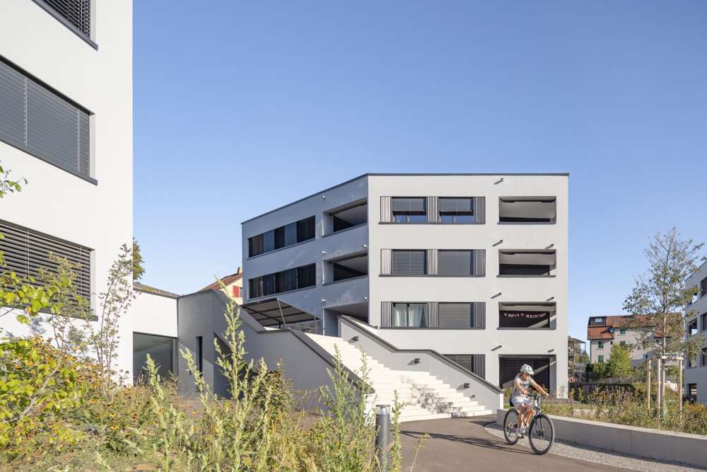 Wohnhof zwischen den Häusern: Freitreppe, Staudenbepflanzung und Wegeführung bilden den gemeinsamen Freiraum; die Loggien sitzen als Einschnitte in der Fassade.