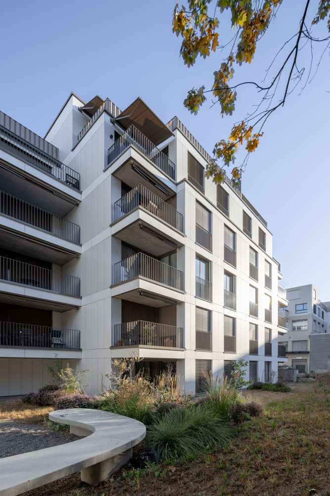 Gebäudeecke zum Hof: vorgelagerte Balkone mit Staketengeländern, Senkrechtmarkisen und Rafflamellenstoren, davor die Sitzbank in Beton.